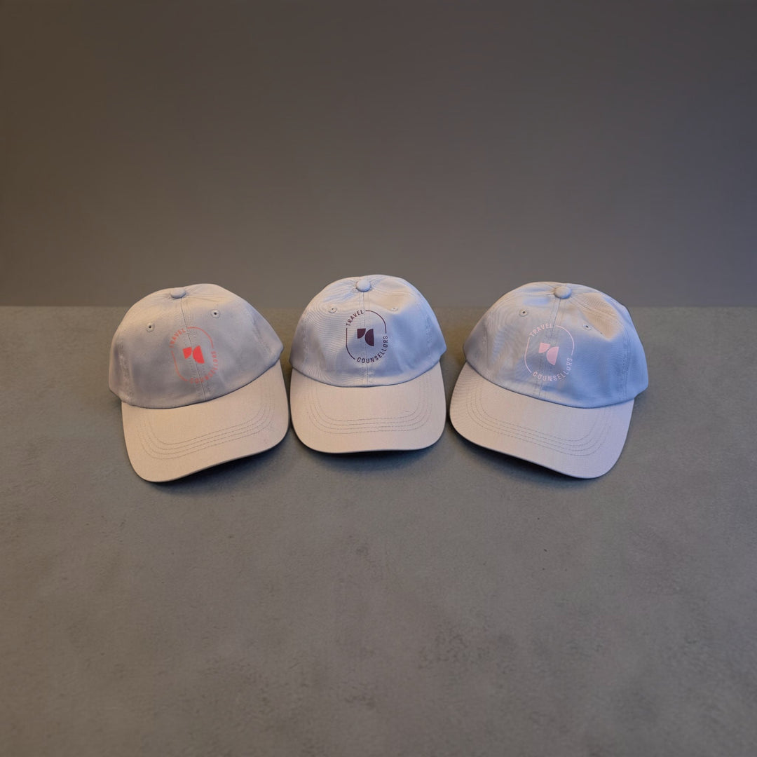 Three Well Travelled Wallets baseball caps in light grey with Travel Counsellors branding on a grey surface.
